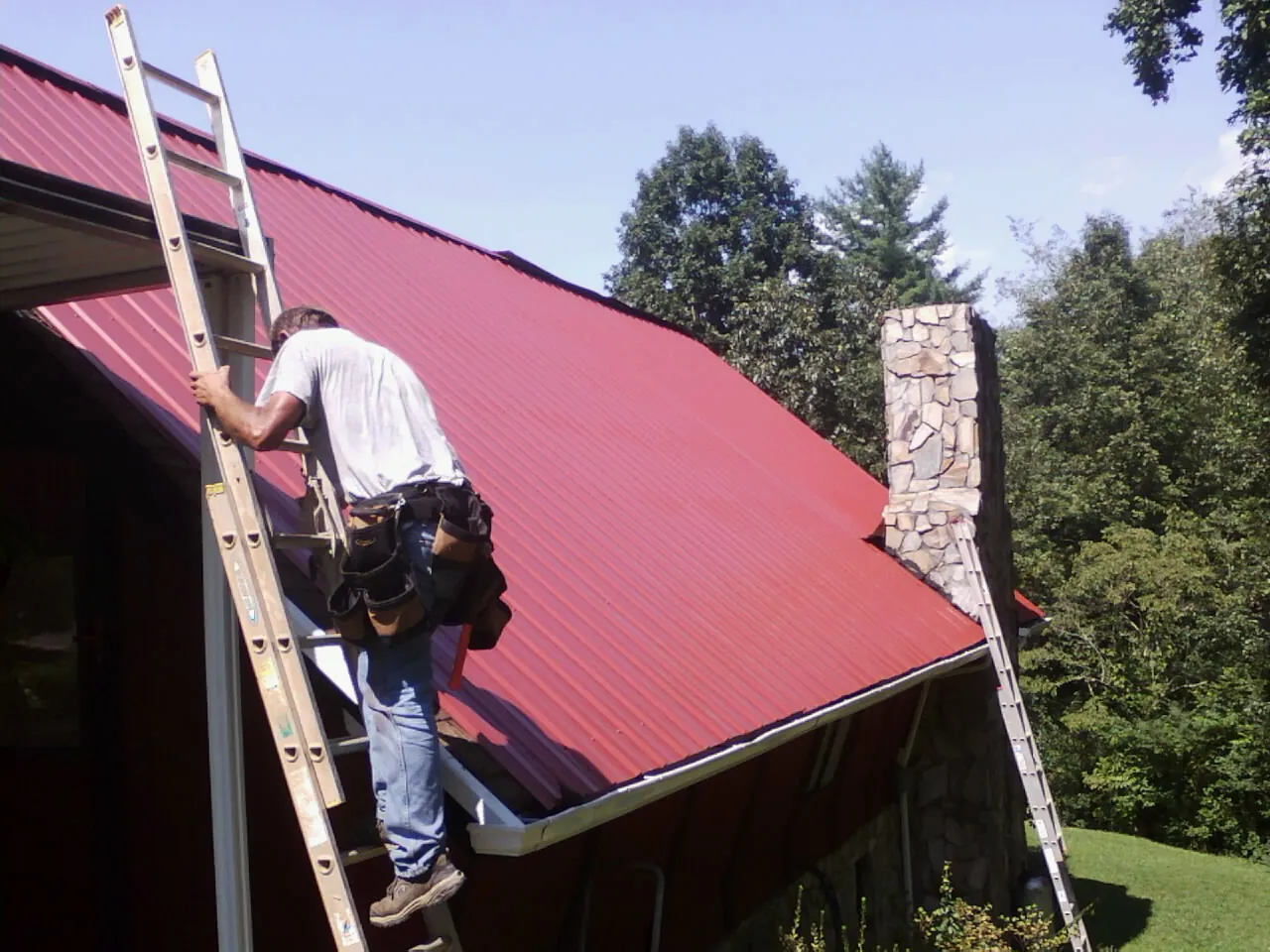 Roofer on a ladder beside a red metal roof and stone chimney — Soffit & Fascia Repair by Roofing Brothers in Pickrell